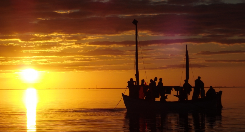 the silhouette of a sailboat holding a group of people is illuminated as the sun sets behind them. The sky appears in vibrant orange.
